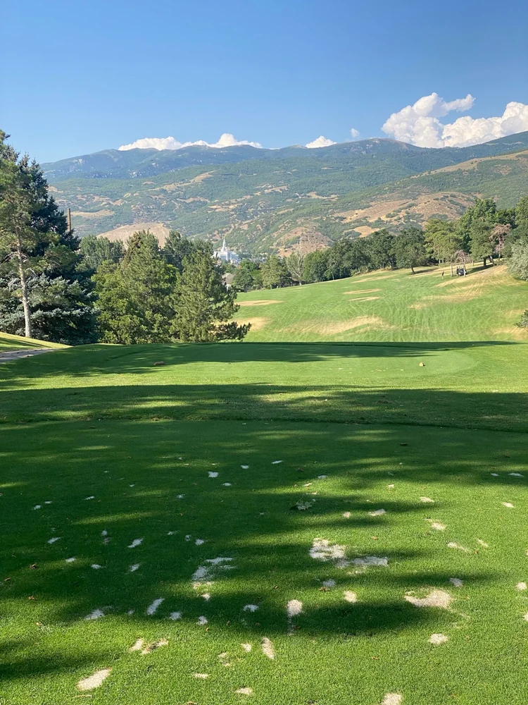 A shaded tee and tree-lined fairway at Bountiful Ridge, with mountain slopes beyond.
