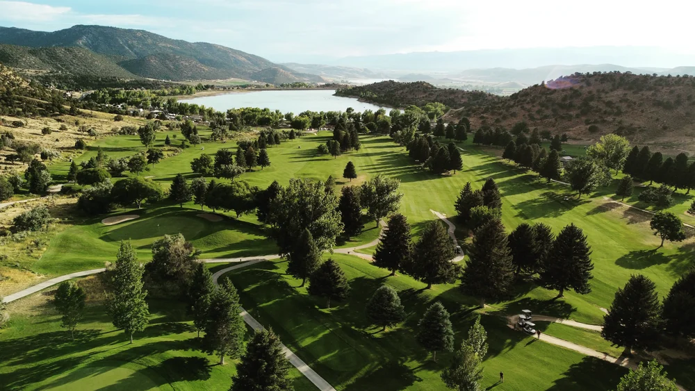 An aerial view of tree-lined fairways, greens and cart paths at Palisade, with a reservoir and hills beyond.