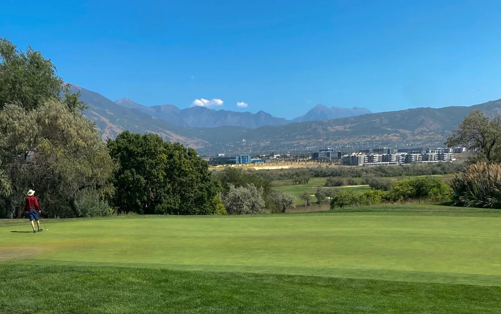 A golfer on a green at Riverbend Golf Course, with trees, buildings and mountain ridges beyond.