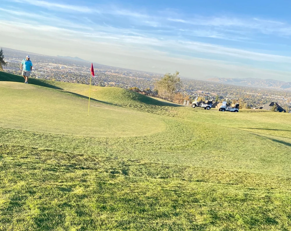 A flag and golfer on a green at South Mountain Golf Course, with carts and the valley beyond.
