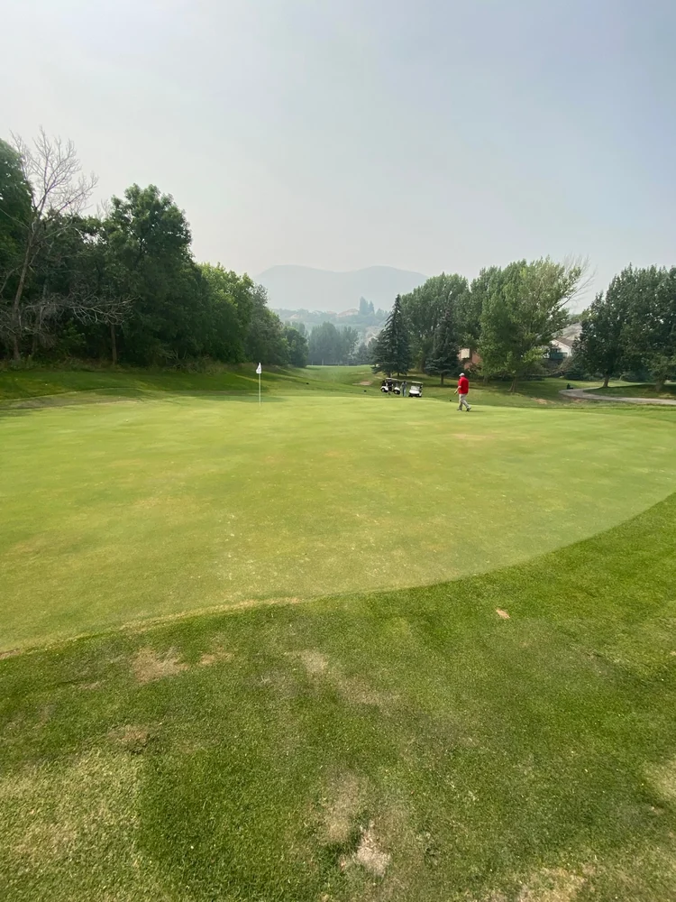 A flag and golfer on a tree-lined green at The Oaks at Spanish Fork, with carts and hills beyond.