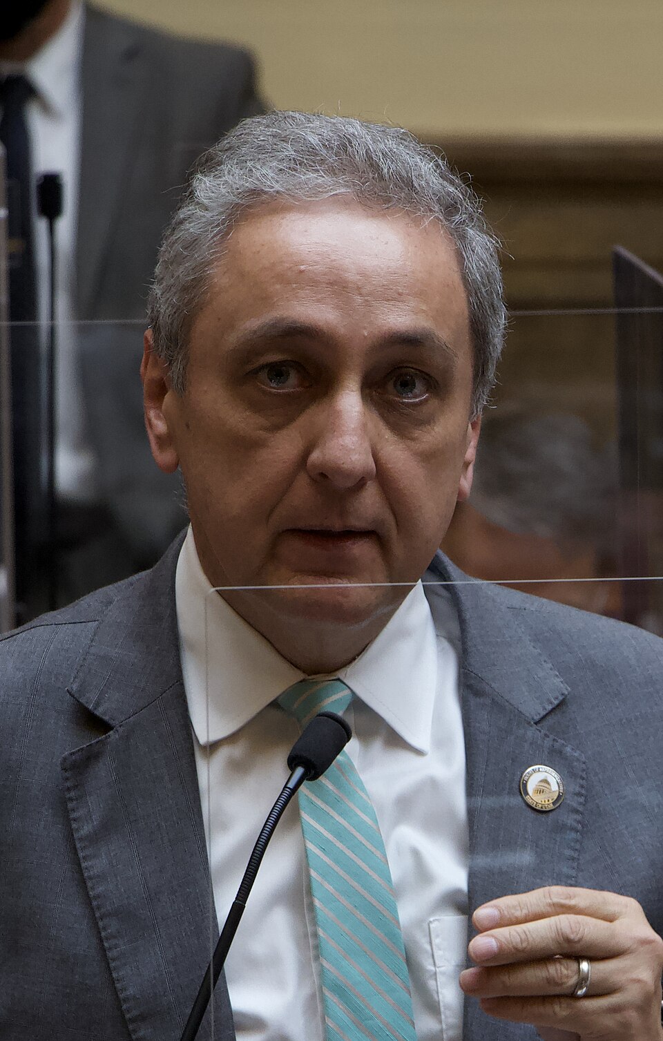 Cory Maloy in a gray suit and turquoise striped tie speaks into a microphone behind a clear divider.