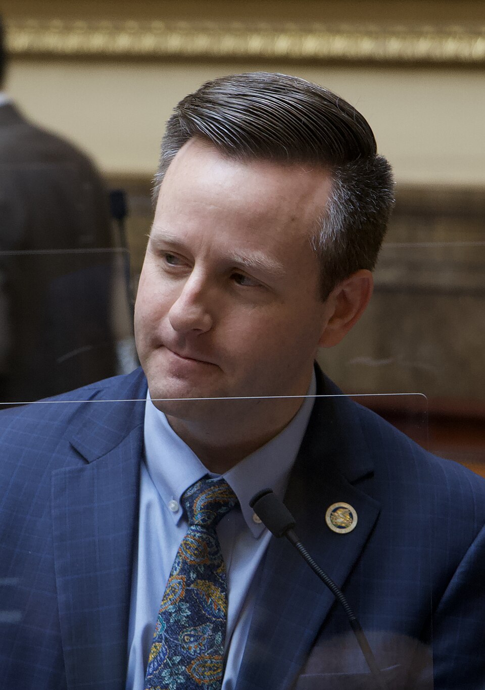 Brady Brammer, wearing a blue suit and patterned tie, sits behind a microphone and clear divider.