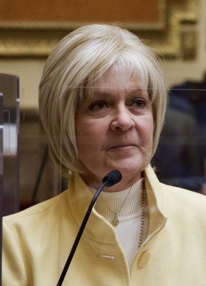 Christine Watkins in a pale yellow jacket at a chamber microphone.