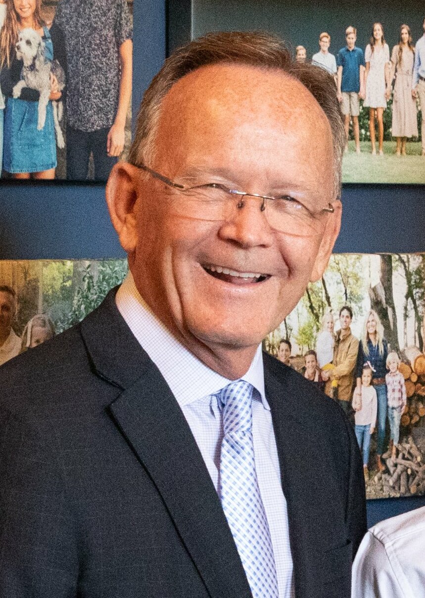 Stuart Adams smiling in glasses, a dark jacket and a light blue tie.