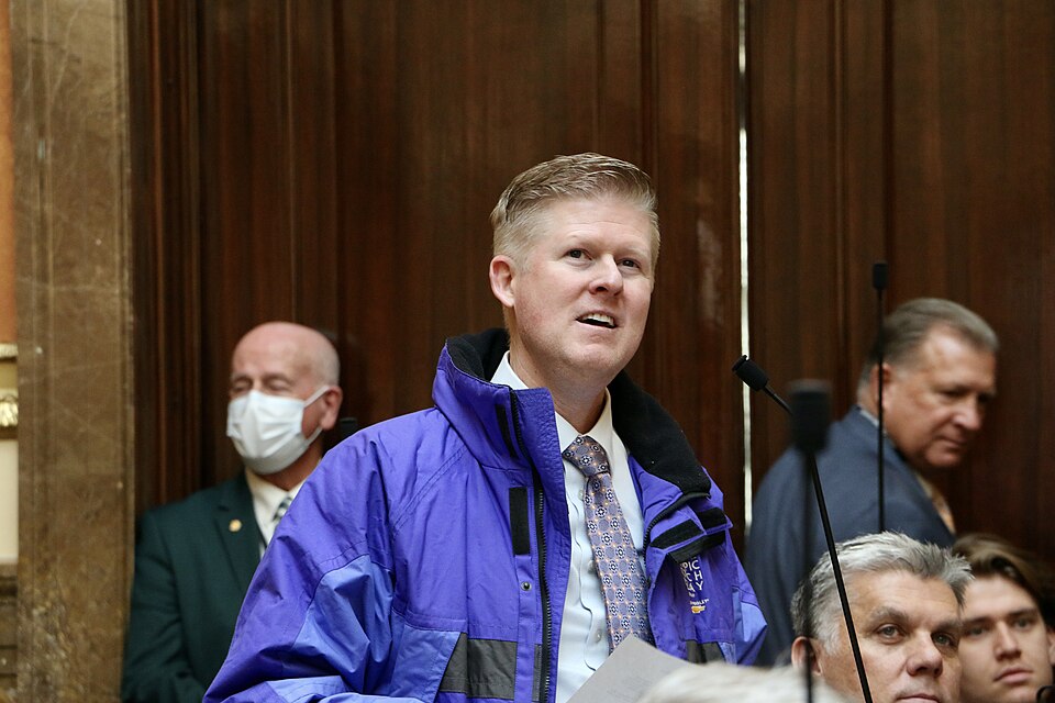 Jon Hawkins, wearing a blue jacket over a shirt and tie, stands at a microphone in the Utah House chamber.