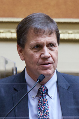 Keven Stratton, wearing a blue suit and patterned tie, speaks at a microphone.