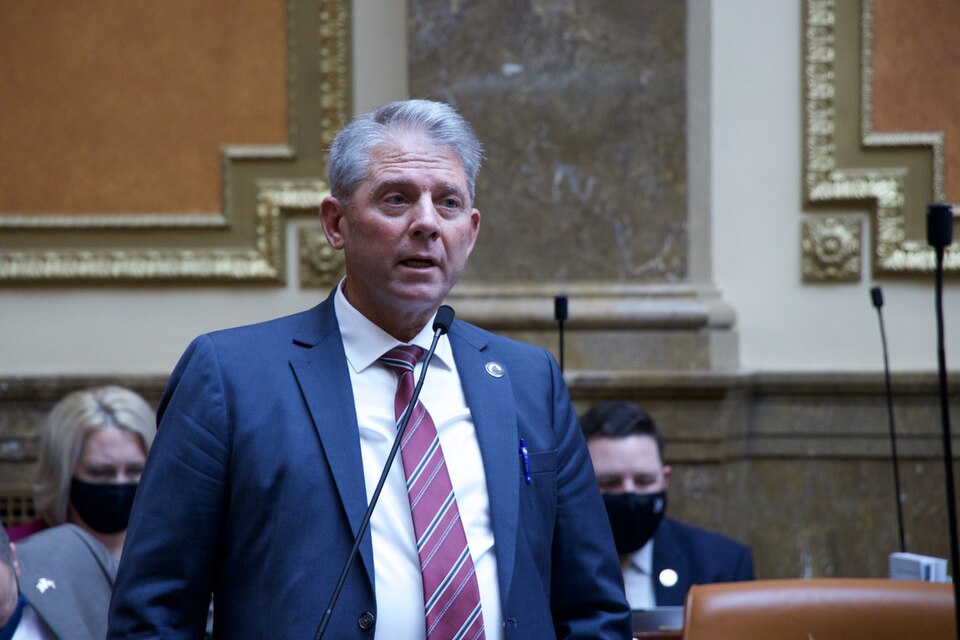 Rex Shipp, wearing a blue suit and red striped tie, stands at a microphone.