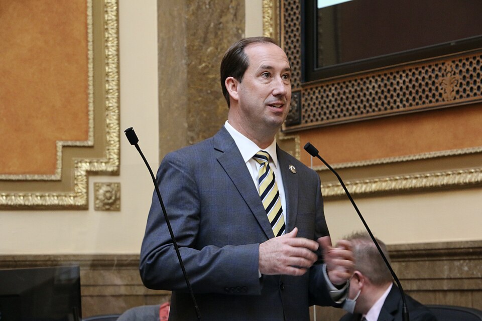 Stephen Whyte, wearing a gray suit and striped tie, stands and gestures beside microphones.