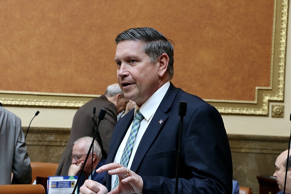 Val Peterson, wearing a dark suit and striped tie, stands and gestures at a microphone.