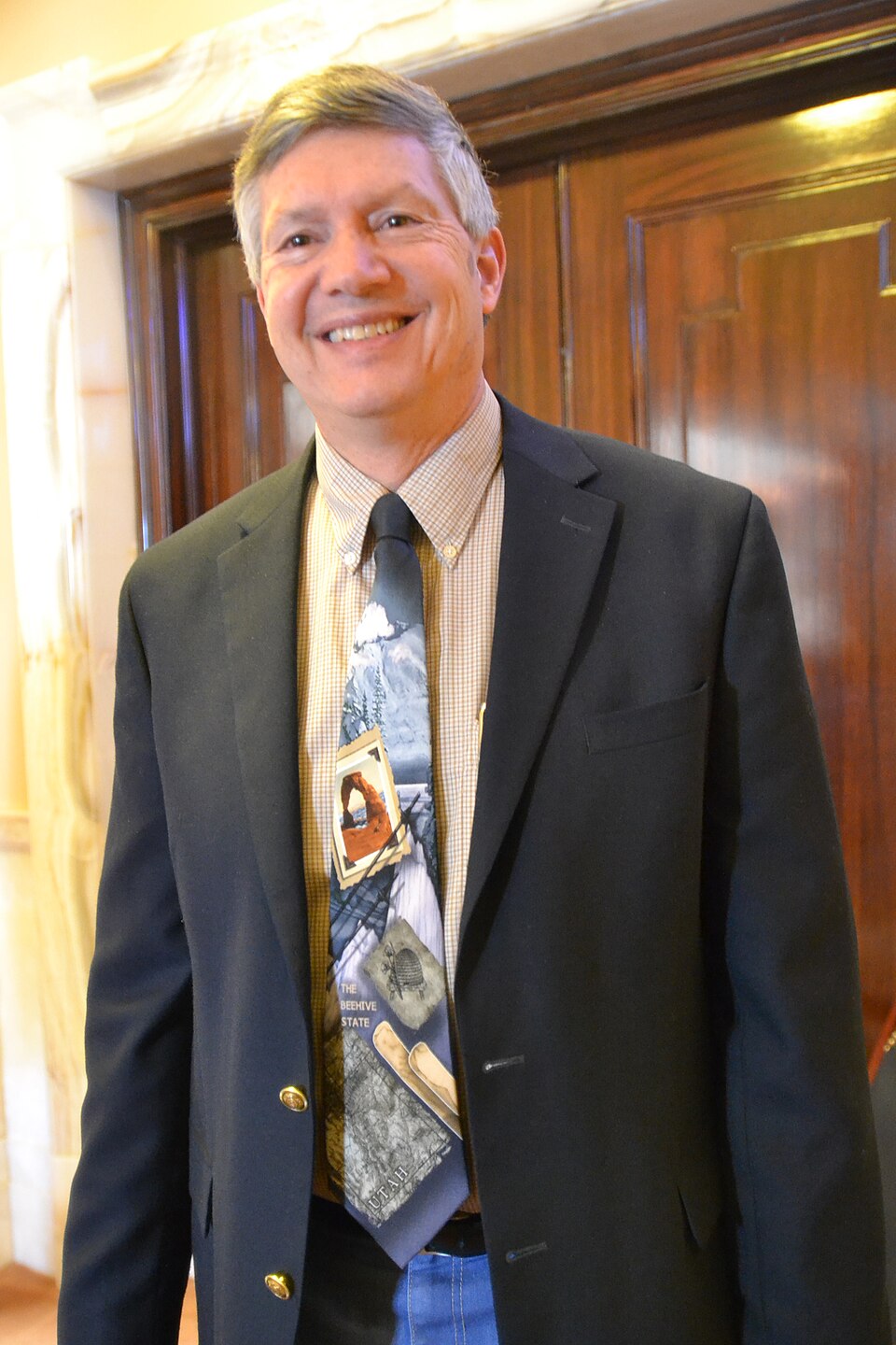 Wayne Harper smiling in a dark jacket and illustrated Utah tie.